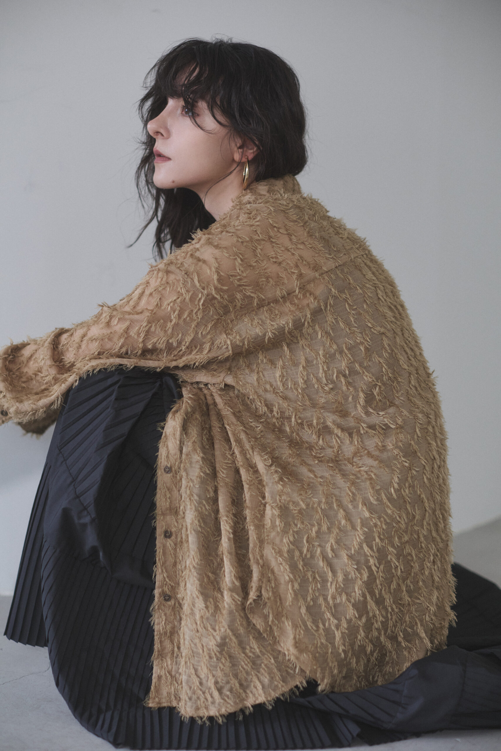 キャメル色のシャギーカーディガンと黒いロングプリーツスカートを着てしゃがんでいる女性モデル写真