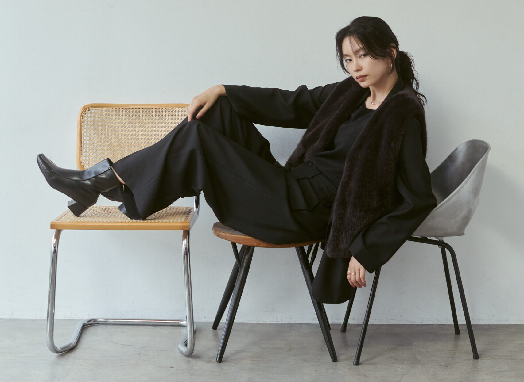 黒いトップス＆パンツにこげ茶のファーベストを着て椅子に座っている女性モデル写真